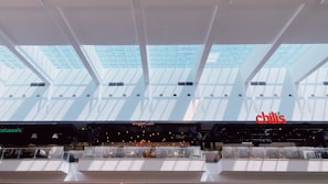 A spacious, modern atrium with skylights allowing natural light to fill the space. Below the glass ceiling are several food establishments including Degas Cafe, and Chili's. The architecture features clean lines and a bright atmosphere due to abundant sunlight.