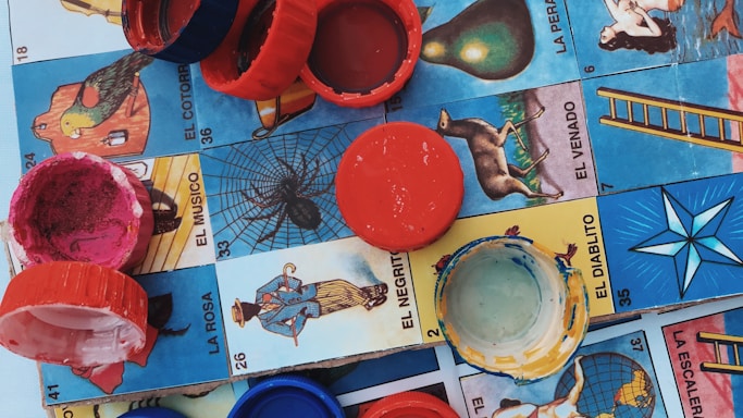 Colorful plastic bottle caps are scattered on a Mexican Lotería board, featuring various illustrated cards with vibrant imagery and Spanish text.