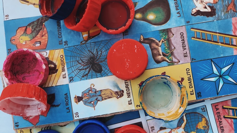 Colorful plastic bottle caps are scattered on a Mexican Lotería board, featuring various illustrated cards with vibrant imagery and Spanish text.