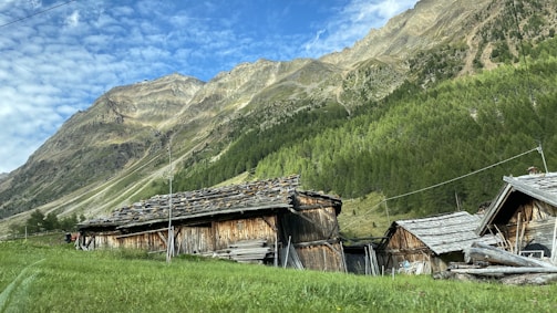Wooden cabins with rustic roofs are situated on a grassy slope, surrounded by a backdrop of towering mountains and lush green forests. The sky is clear with some scattered clouds, adding depth to the picturesque landscape.