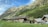 Wooden cabins with rustic roofs are situated on a grassy slope, surrounded by a backdrop of towering mountains and lush green forests. The sky is clear with some scattered clouds, adding depth to the picturesque landscape.