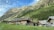 Wooden cabins with rustic roofs are situated on a grassy slope, surrounded by a backdrop of towering mountains and lush green forests. The sky is clear with some scattered clouds, adding depth to the picturesque landscape.