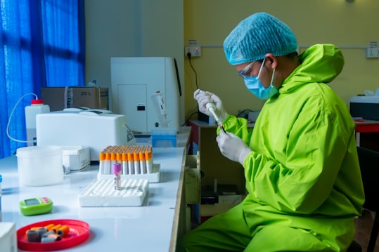 A person in protective attire, including a bright green suit, gloves, a face mask, and a hair cap, is working in a laboratory setting. They are holding a syringe and appear to be performing some tests or experiments. Numerous test tubes, some with red caps and filled with liquid, are organized on a table. The room has scientific equipment, including a centrifuge, and blue curtains allow natural light to filter through the window.