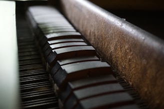 A close-up view of a section of a piano, focusing on the hammers and strings inside the piano. The perspective is angled, showing the texture of the wood and the alignment of the hammers in a dimly lit environment.