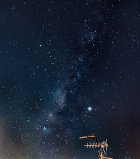 A night sky filled with countless stars and a band of the Milky Way galaxy stretching diagonally across. In the lower right corner, an antenna structure is silhouetted against the starry background.
