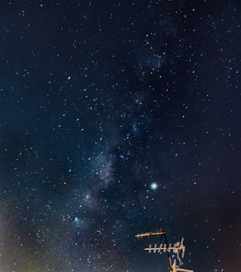 A night sky filled with countless stars and a band of the Milky Way galaxy stretching diagonally across. In the lower right corner, an antenna structure is silhouetted against the starry background.