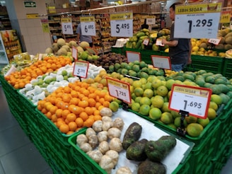 A market display featuring a variety of fresh fruits such as oranges, green apples, avocados, and melons, arranged neatly in green plastic crates. There are multiple price tags shown prominently above the fruits. A shopper is browsing the produce in the background.