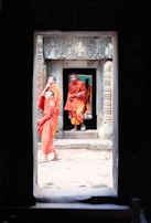 A group of young monks wearing orange robes stands in an ancient stone doorway. The walls are intricately carved with detailed patterns, and the scene is framed by a dark archway, creating a sense of depth.