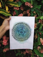 A person is holding a piece of paper with a detailed mandala design that transitions from dark blue in the center to light green at the edges. The background consists of lush green foliage and pink flowers.