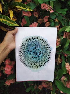A person is holding a piece of paper with a detailed mandala design that transitions from dark blue in the center to light green at the edges. The background consists of lush green foliage and pink flowers.