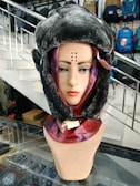 A mannequin head is adorned with a faux fur hat and a colorful scarf. The setting is indoors, likely a store with various bags on display in the background. A staircase is visible, as well as glass shelves displaying small items.