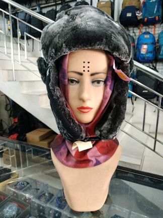 A mannequin head is adorned with a faux fur hat and a colorful scarf. The setting is indoors, likely a store with various bags on display in the background. A staircase is visible, as well as glass shelves displaying small items.