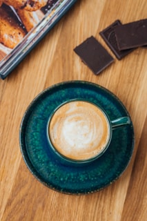 A cup of coffee is placed on a deep green saucer, sitting on a wooden surface. Two pieces of dark chocolate are positioned next to an open magazine displaying a picture of pastries.