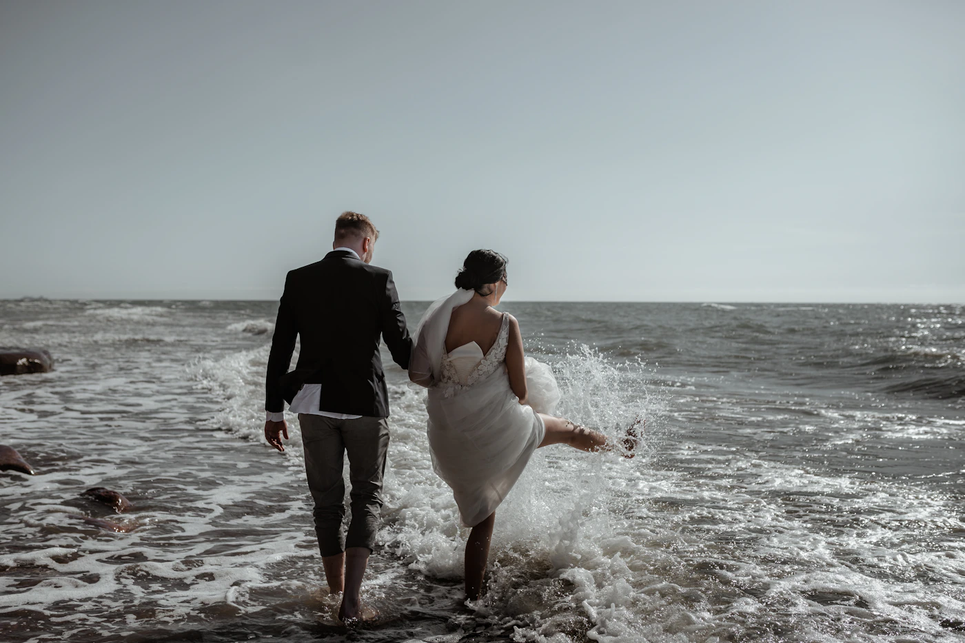 Groom in dark suit holding bride in white dress at the edge of a rocky ocean coastline during daytime