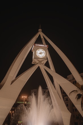 A tall, modern clock tower with a geometric design and the Omega branding is prominently featured. The structure is illuminated against a night sky, with a fountain spraying water in the foreground. Surrounding buildings and palm trees can be seen, capturing a cityscape atmosphere.