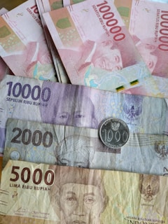 Various denominations of Indonesian Rupiah currency notes are scattered, including 100,000, 10,000, 5,000, and 2,000 Rupiah notes, along with a 1,000 Rupiah coin. The notes display intricate designs and portraits, showcasing notable figures. The arrangement emphasizes the diverse colors and sizes of the currency.