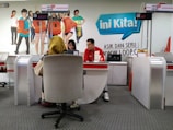 A customer service area features a desk with two employees, one wearing a red uniform and the other assisting a customer. The setting includes a colorful backdrop with life-sized cutouts of young people and the word 'Loop' in large letters. The counter displays digital numbers, and promotional materials are placed on the desk.