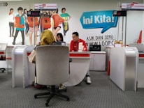 A customer service area features a desk with two employees, one wearing a red uniform and the other assisting a customer. The setting includes a colorful backdrop with life-sized cutouts of young people and the word 'Loop' in large letters. The counter displays digital numbers, and promotional materials are placed on the desk.