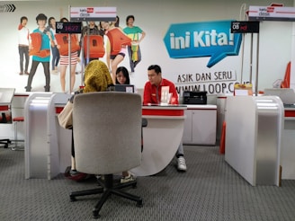 A customer service area features a desk with two employees, one wearing a red uniform and the other assisting a customer. The setting includes a colorful backdrop with life-sized cutouts of young people and the word 'Loop' in large letters. The counter displays digital numbers, and promotional materials are placed on the desk.