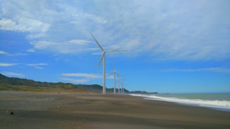 Several wind turbines are lined up along a beach with a clear blue sky and scattered clouds above. The shoreline stretches to the right with waves gently crashing onto the sand. Green hills are visible in the distance, complementing the coastal landscape.