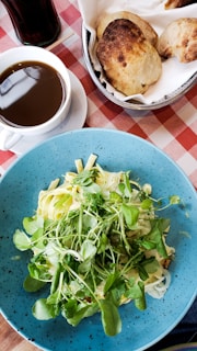 A blue plate filled with a fresh salad topped with leafy greens is situated on a table with a red and white checkered tablecloth. Next to it, there is a cup of black coffee placed on a saucer, and a basket containing two pieces of rustic bread, showing a golden-brown crust.