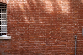 A red brick wall with visible mortar between the bricks. To the left, there is a window with metal bars covering it and a stone sill. A small metal pipe or conduit runs vertically near the right edge of the image.