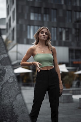 A woman stands confidently in an urban setting, wearing a light green sleeveless top and black pants. Her pose is relaxed and stylish, with her hand on her hip. The background features modern, dark-toned architectural structures and blurred outdoor umbrellas, suggesting a city environment.