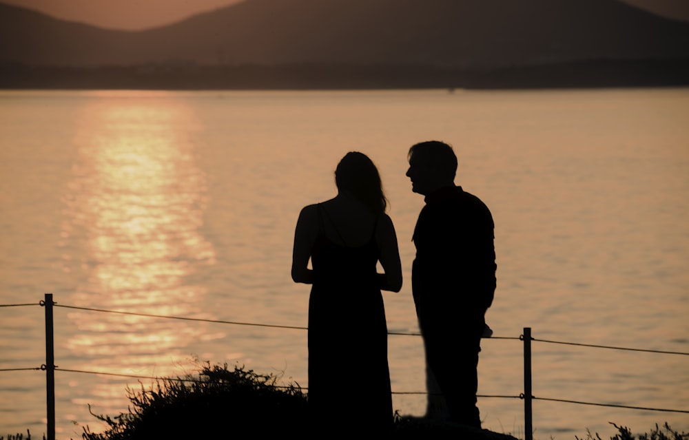 silhouette of man and woman standing near body of water during sunset