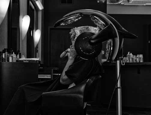 A person sits in a salon chair under a hair drying machine, with their hair wrapped in foil. The setting appears modern, with various hair products visible on a countertop. The lighting is soft and focused, enhancing the black-and-white aesthetic of the image.