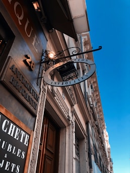 A street view of a building facade with ornate architectural details and a wooden door. A vintage-style sign hangs above, featuring the word 'Antiquities'. The signage and facade details, including the number 28, suggest a store or shop entrance. The sky is clear and blue, adding contrast to the historic aesthetic of the building.