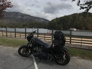 A motorcycle is parked on a paved area near a body of water, with mountains and trees in the background. The sky is cloudy, and there is a wooden fence separating the road from the water. The motorcycle is black and has a large backpack attached, along with a bottle of Coca-Cola visible.