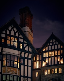 A Tudor-style building with intricate black and white timber framing is illuminated at night. Warm light glows through multiple rectangular windows, and a tall red brick chimney rises prominently against the dark evening sky.
