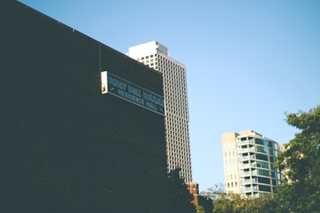 Urban scene featuring a dark building wall with a sign that reads 'Moody Bible Institute Residence Hall.' In the background, several high-rise buildings are visible under a clear blue sky, with some lush greenery to the side.