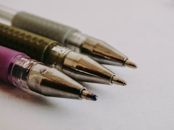 A close-up of four pen tips aligned diagonally on a light background. The pens have metallic tips, and the barrels are partially visible with different colors including purple and gray. The focus is on the pen tips, highlighting their precision and shine.