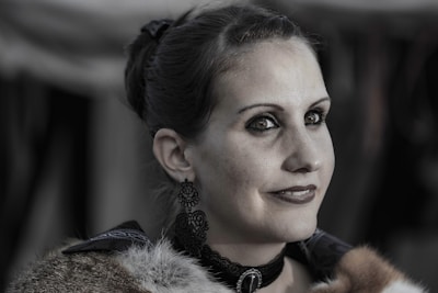 A woman with dark hair styled in an updo is wearing elaborate earrings and a choker necklace. Her attire features fur details, and she has a subtle smile on her face. The background is blurred, putting emphasis on her facial features and attire.