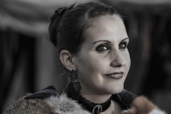 A woman with dark hair styled in an updo is wearing elaborate earrings and a choker necklace. Her attire features fur details, and she has a subtle smile on her face. The background is blurred, putting emphasis on her facial features and attire.
