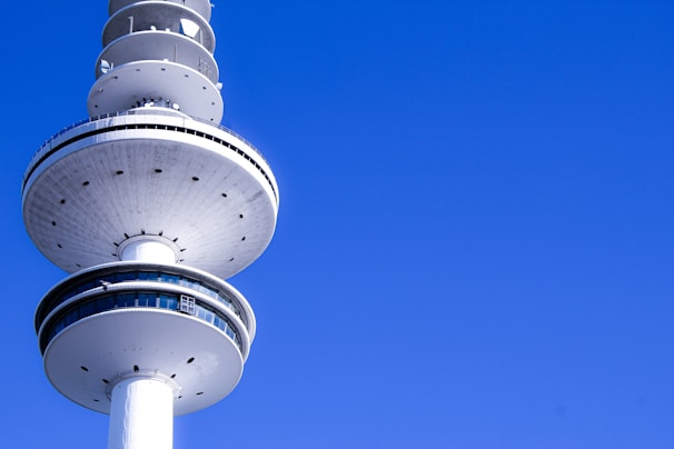 A modern telecommunications tower with a rounded observation deck and several antennae above it, set against a clear blue sky. The structure has a sleek design with a prominent white and silver coloration, and features windows around the deck.