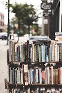 A display of various books arranged on shelves set up outdoors. The books cover a wide range of topics and genres, with titles visible on the spines. The background has a slightly blurred city sidewalk and storefronts, indicative of an urban street setting.