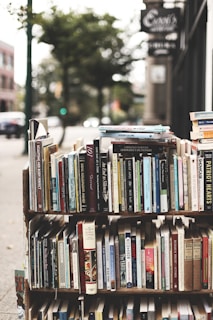 A display of various books arranged on shelves set up outdoors. The books cover a wide range of topics and genres, with titles visible on the spines. The background has a slightly blurred city sidewalk and storefronts, indicative of an urban street setting.