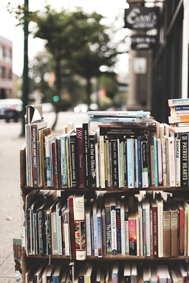 A display of various books arranged on shelves set up outdoors. The books cover a wide range of topics and genres, with titles visible on the spines. The background has a slightly blurred city sidewalk and storefronts, indicative of an urban street setting.