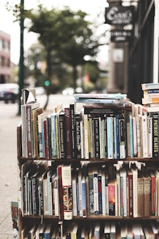 A display of various books arranged on shelves set up outdoors. The books cover a wide range of topics and genres, with titles visible on the spines. The background has a slightly blurred city sidewalk and storefronts, indicative of an urban street setting.