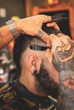A person with tattoos on their hands is giving a haircut using scissors and a comb. The focus is on the close-up detail of the haircut process, highlighting the person's ear with a tunnel plug and the stylist's tattoos.