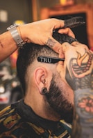 A person with tattoos on their hands is giving a haircut using scissors and a comb. The focus is on the close-up detail of the haircut process, highlighting the person's ear with a tunnel plug and the stylist's tattoos.