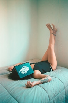A person is lying on a bed with their legs raised against a wall. A book is placed open on their face, and a tattoo is visible on their arm. The room has minimal decoration and soft lighting.