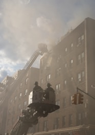 Firefighters are in a bucket lift extended towards a smoke-filled multi-story building. The scene is hazy due to the smoke, and the building has a brick facade. A traffic light is visible in the foreground, suggesting an urban environment.