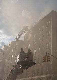 Firefighters are in a bucket lift extended towards a smoke-filled multi-story building. The scene is hazy due to the smoke, and the building has a brick facade. A traffic light is visible in the foreground, suggesting an urban environment.