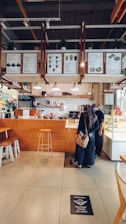 The interior of a cozy café with a wooden counter and a menu displayed overhead. A person wearing a long dress and headscarf is standing at the counter, possibly ordering or interacting with the barista. A bar stool and some seating are visible, along with decorative plants and a well-organized counter area.
