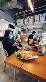 A cozy caf&eacute; setting with a focus on a wooden table featuring a meal with chips and greens, alongside a cup of coffee, dessert, and orange drink. Two individuals sitting in the caf&eacute; are wearing masks, indicating a concern for health safety. The interior has a modern design with menu boards hanging from the ceiling and soft lighting.