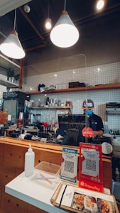 A small coffee shop counter featuring hanging lights and a barista standing behind a glass partition. Shelves with coffee supplies and knick-knacks adorn the tiled wall. Two QR code payment options are displayed prominently on the counter. A bottle of sanitizer and menu flyers are placed next to the payment display.