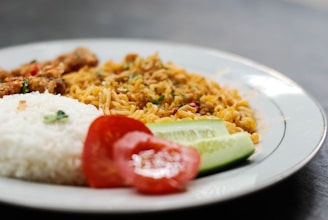 A plate with a serving of white rice, fried noodles, a slice of tomato, and a piece of cucumber. The noodles appear well-cooked with sprinkled herbs and small pieces of meat, adding color and texture. The meal is arranged neatly on the plate.
